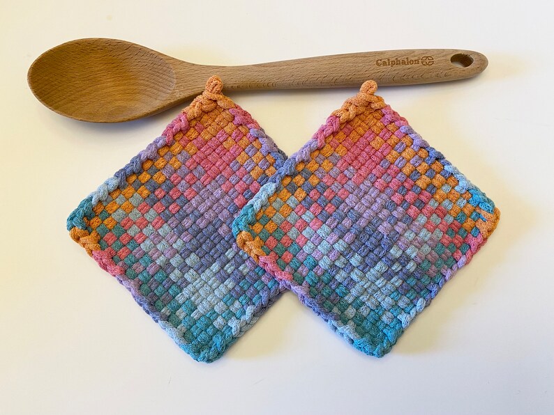 May include: Two square, woven potholders in shades of blue, pink, orange, and purple. A wooden spoon rests above them. The potholders have a braided edge and a loop for hanging. The spoon is light brown and has the word "Calphalon" printed on it.