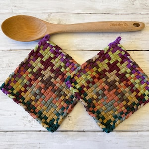 May include: Two square woven potholders featuring a multicolored checkered pattern in shades of purple, green, orange, and yellow, positioned below a light brown wooden spoon with the brand name Calphalon engraved on the handle.