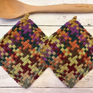 May include: Two square potholders featuring a woven, multicolored pattern in shades of purple, green, orange, and brown, positioned below a light-colored wooden spoon with the brand name Calphalon engraved on the handle.