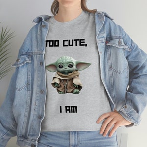 May include: A light gray t-shirt featuring a graphic of a green alien character with large ears and the text "TOO CUTE, I AM" in black block letters. The alien is wearing a brown robe. The person is wearing a denim jacket.