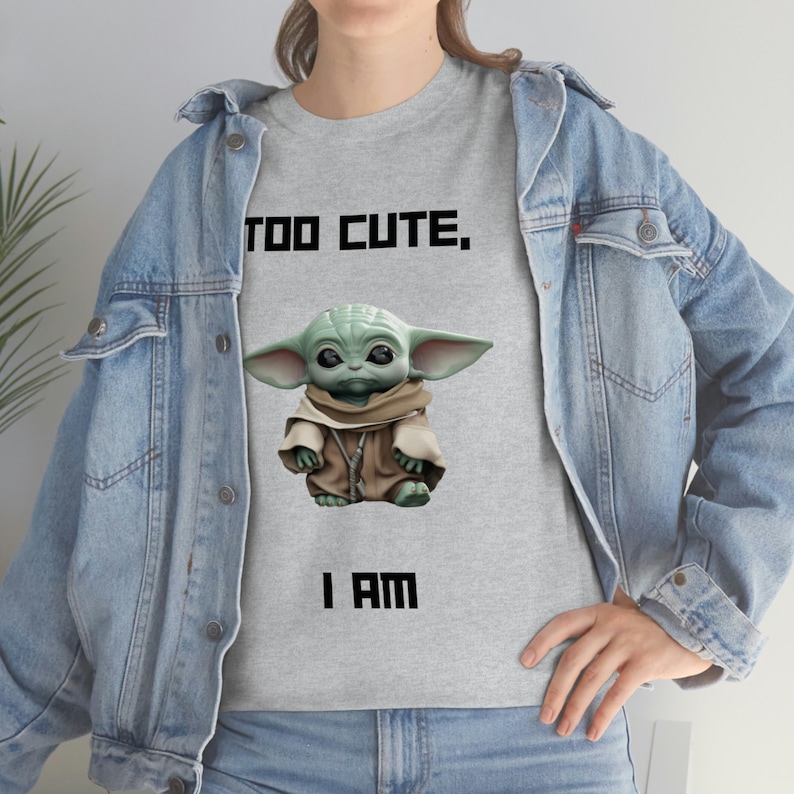 May include: A light gray t-shirt featuring a graphic of a green alien character with large ears and the text "TOO CUTE, I AM" in black block letters. The alien is wearing a brown robe. The person is wearing a denim jacket.
