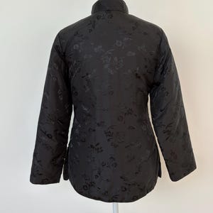 May include: Black quilted jacket with a mandarin collar and long sleeves. The jacket features a subtle floral pattern throughout the fabric. The garment is displayed on a white mannequin, showcasing its design and fit. A classic piece for layering.
