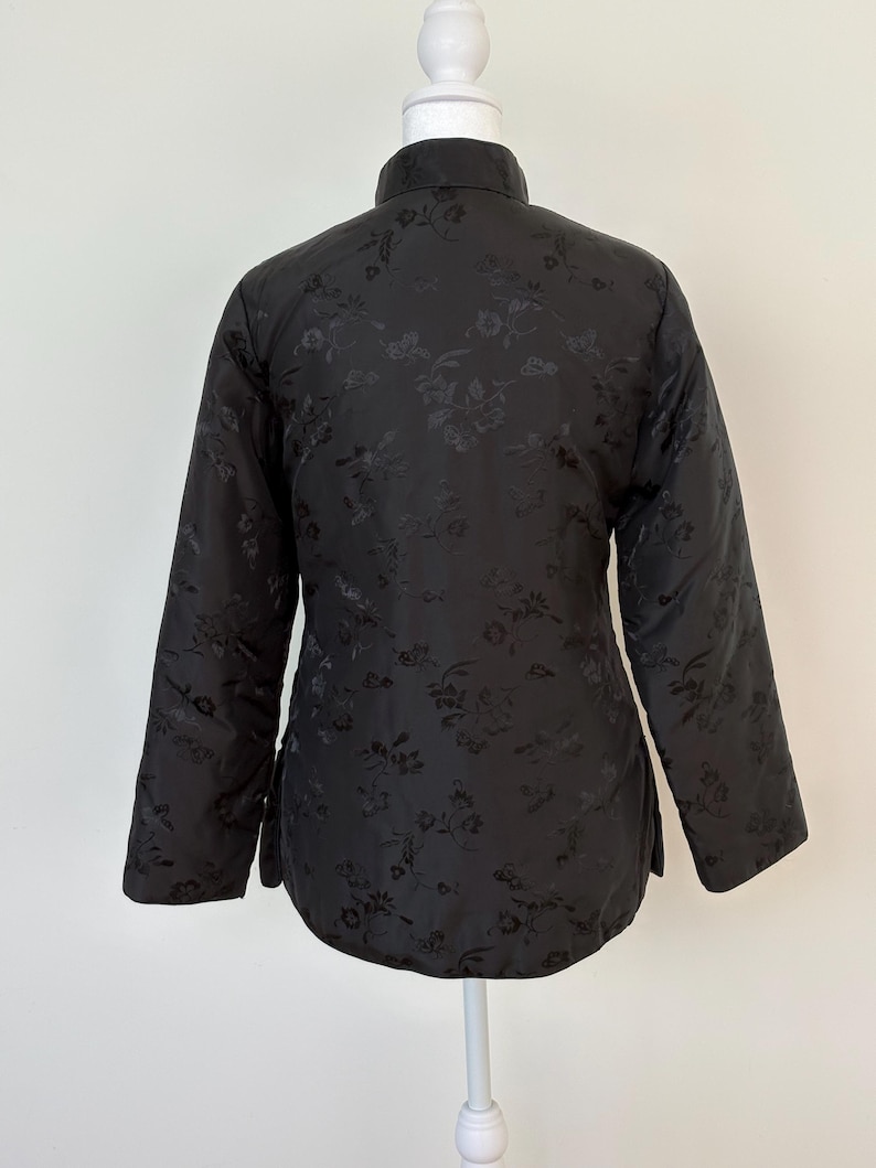 May include: Black quilted jacket with a mandarin collar and long sleeves. The jacket features a subtle floral pattern throughout the fabric. The garment is displayed on a white mannequin, showcasing its design and fit. A classic piece for layering.