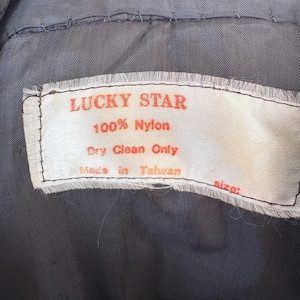 May include: Close-up of a tag from a garment. The tag is white with red text that reads "LUCKY STAR" and "100% Nylon." Below the text is "Dry Clean Only" and "Made in Taiwan." The background is a dark gray fabric.