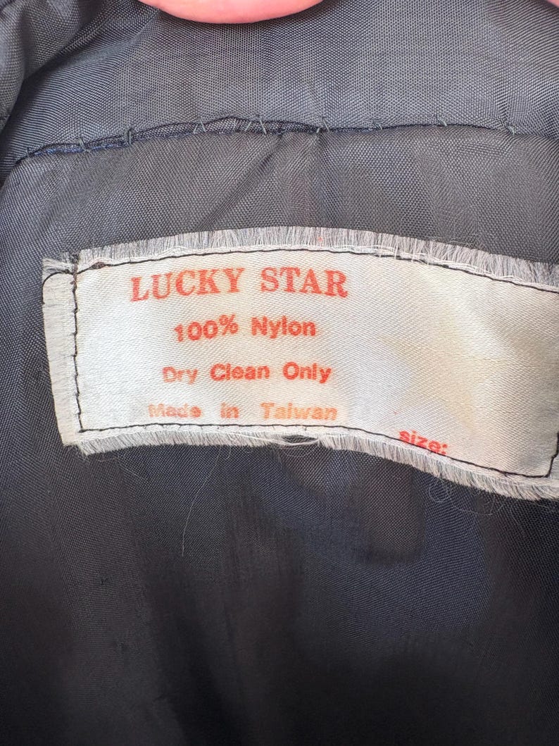 May include: Close-up of a tag from a garment. The tag is white with red text that reads "LUCKY STAR" and "100% Nylon." Below the text is "Dry Clean Only" and "Made in Taiwan." The background is a dark gray fabric.