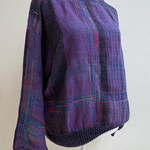 May include: A purple plaid sweater with a high neck and long sleeves. The sweater features a ribbed collar, cuffs, and hem in a dark purple color. The plaid pattern includes lines of red, green, and yellow.