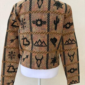 K&ouml;nnte beinhalten: Eine braune, beige und schwarze Jacke mit einem Muster aus s&uuml;dwestlichen Symbolen. Das Design umfasst geometrische Formen, Kakteen und andere vom W&uuml;stenstil inspirierte Motive. Die Jacke wird auf einer wei&szlig;en Schaufensterpuppe pr&auml;sentiert.