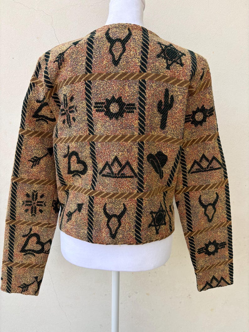 K&ouml;nnte beinhalten: Eine braune, beige und schwarze Jacke mit einem Muster aus s&uuml;dwestlichen Symbolen. Das Design umfasst geometrische Formen, Kakteen und andere vom W&uuml;stenstil inspirierte Motive. Die Jacke wird auf einer wei&szlig;en Schaufensterpuppe pr&auml;sentiert.