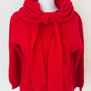 Puede incluir: Un suéter rojo brillante con una textura de punto acanalado. El suéter presenta un cuello alto, tipo bufanda, que se ata con un lazo en la parte delantera. Las mangas largas y los bolsillos completan el diseño. Una prenda clásica y cálida.