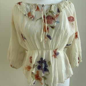 May include: Cream-coloured blouse with floral embroidery. The blouse has a ruffled neckline, three-quarter sleeves, and a gathered waist. The floral design includes shades of red, orange, purple, and green. The blouse is made of a lightweight, textured fabric.