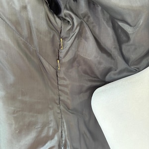 May include: Close-up of the inside lining of a dark gray jacket. The lining is a smooth, shiny fabric with visible stitching and gold-colored pins holding the fabric together. A white mannequin shoulder is visible on the right side.