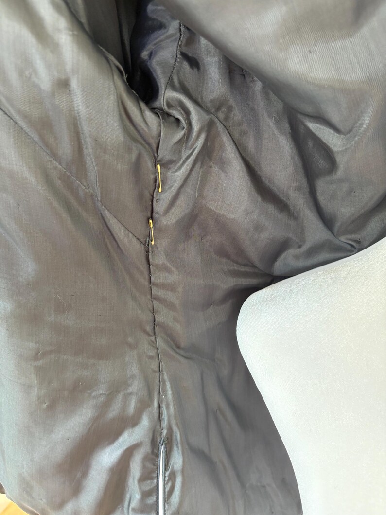 May include: Close-up of the inside lining of a dark gray jacket. The lining is a smooth, shiny fabric with visible stitching and gold-colored pins holding the fabric together. A white mannequin shoulder is visible on the right side.