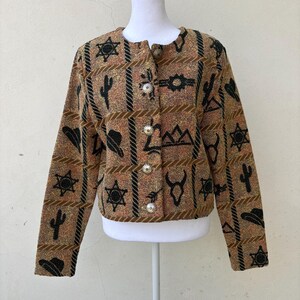 K&ouml;nnte beinhalten: Eine braun, beige und schwarz gemusterte Jacke mit Western-Motiv. Die Jacke hat einen runden Kragen, lange &Auml;rmel und silberne Kn&ouml;pfe. Das Muster umfasst Sterne, Kakteen, H&uuml;te und andere vom Western inspirierte Motive.
