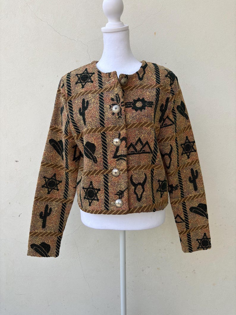 K&ouml;nnte beinhalten: Eine braun, beige und schwarz gemusterte Jacke mit Western-Motiv. Die Jacke hat einen runden Kragen, lange &Auml;rmel und silberne Kn&ouml;pfe. Das Muster umfasst Sterne, Kakteen, H&uuml;te und andere vom Western inspirierte Motive.