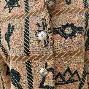 K&ouml;nnte beinhalten: Eine gemusterte Jacke in Beige, Braun und Gr&uuml;n mit silberfarbenen Kn&ouml;pfen. Die Jacke zeigt ein s&uuml;dwestliches Design mit Kakteen, Sternen und geometrischen Formen. Die Kn&ouml;pfe haben ein Blumenmuster.