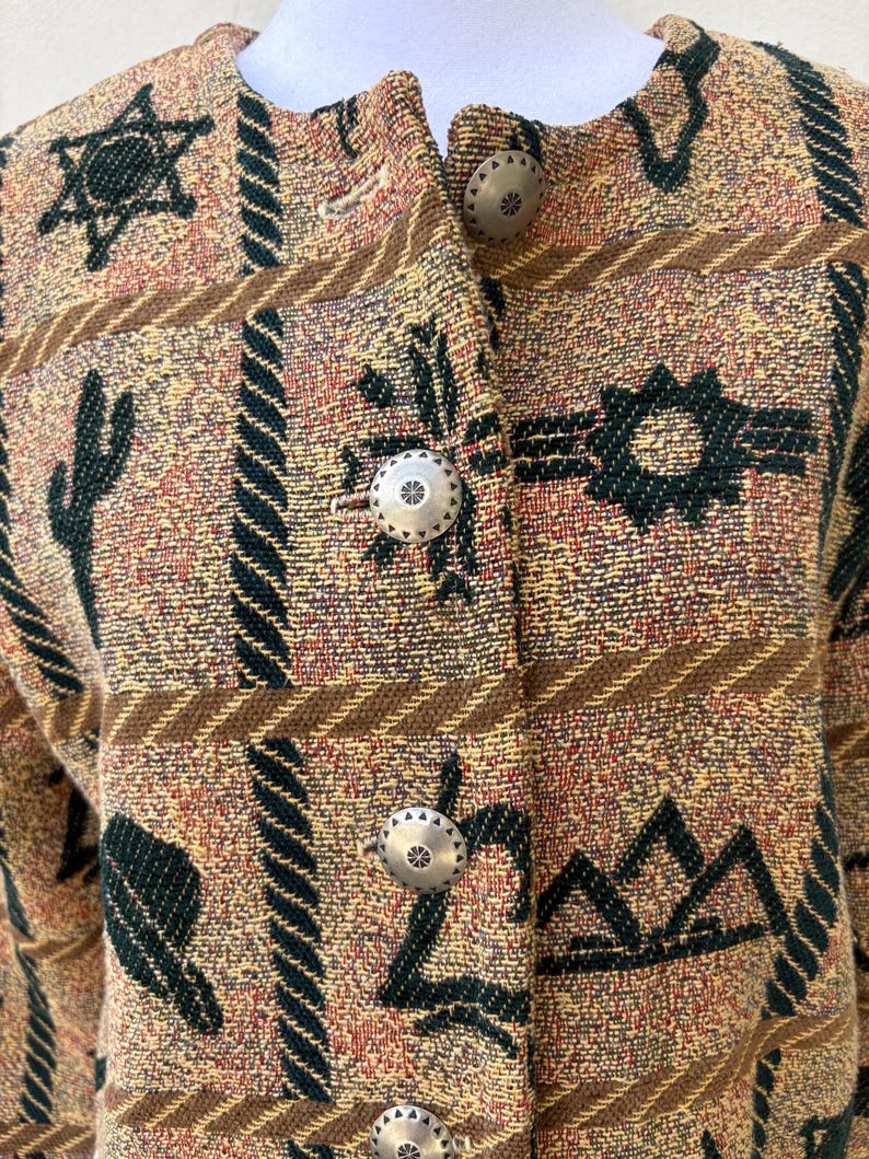 K&ouml;nnte beinhalten: Eine gemusterte Jacke in Beige, Braun und Gr&uuml;n mit silberfarbenen Kn&ouml;pfen. Die Jacke zeigt ein s&uuml;dwestliches Design mit Kakteen, Sternen und geometrischen Formen. Die Kn&ouml;pfe haben ein Blumenmuster.