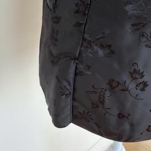May include: A close-up of a black jacket with a floral pattern. The jacket has a subtle sheen and a tailored fit. The floral design is embossed into the fabric, creating a textured effect. The jacket is displayed on a white mannequin.