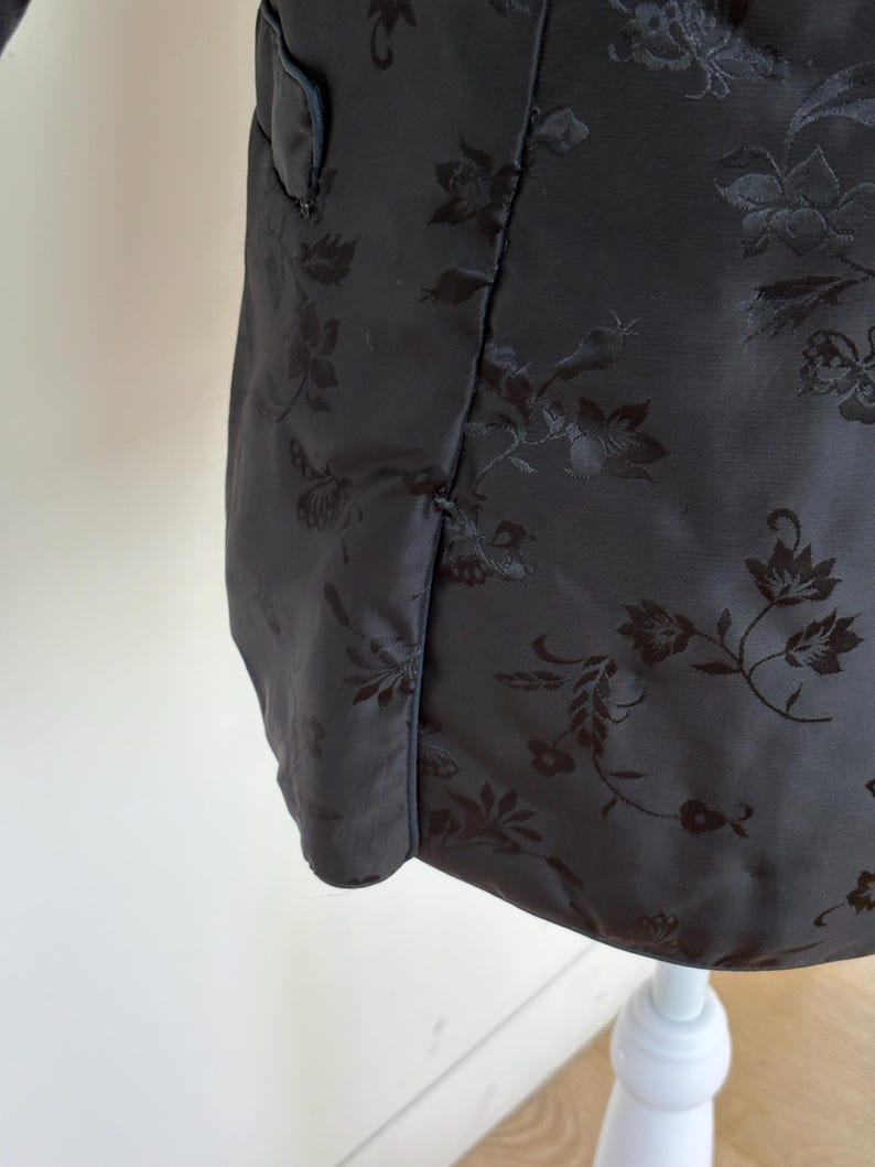 May include: A close-up of a black jacket with a floral pattern. The jacket has a subtle sheen and a tailored fit. The floral design is embossed into the fabric, creating a textured effect. The jacket is displayed on a white mannequin.