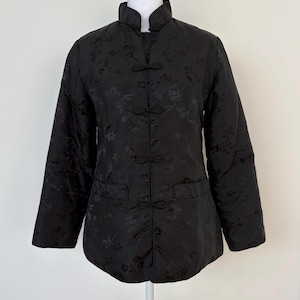 Puede incluir: Chaqueta acolchada negra con cuello mandarín y cierres de rana. La chaqueta presenta un estampado floral y dos bolsillos delanteros. Las mangas son largas y la chaqueta se exhibe en un maniquí.