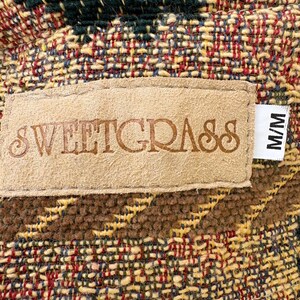 K&ouml;nnte beinhalten: Nahaufnahme eines gewebten Stoffes mit einem Etikett. Der Stoff weist eine Mischung aus Farben auf, darunter Gr&uuml;n, Rot, Gelb und Braun. Auf dem Etikett steht "SWEETGRASS" in gepr&auml;gten Buchstaben. Ein Etikett mit "M/M" ist angebracht.