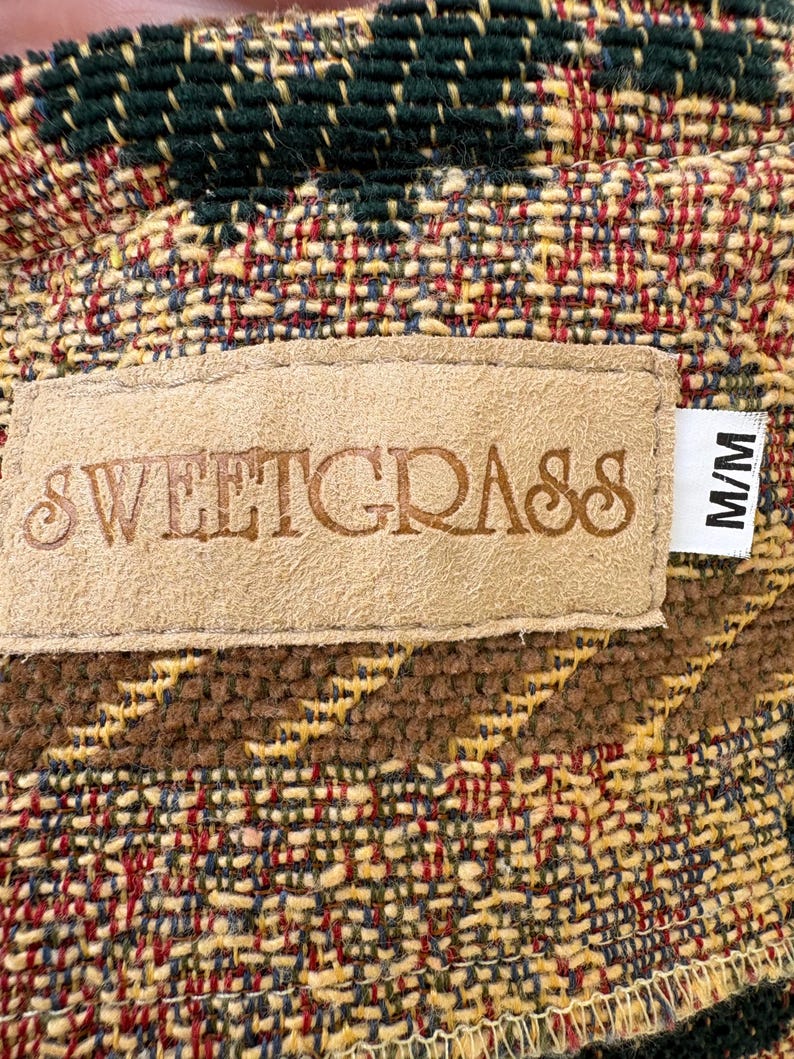 K&ouml;nnte beinhalten: Nahaufnahme eines gewebten Stoffes mit einem Etikett. Der Stoff weist eine Mischung aus Farben auf, darunter Gr&uuml;n, Rot, Gelb und Braun. Auf dem Etikett steht "SWEETGRASS" in gepr&auml;gten Buchstaben. Ein Etikett mit "M/M" ist angebracht.