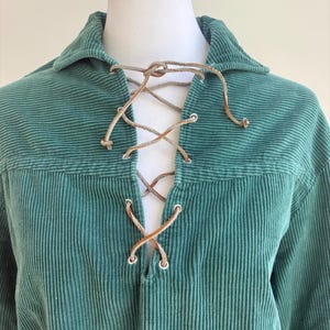 May include: A teal corduroy shirt featuring a collared neckline and a front placket with silver-toned metal eyelets. The placket is laced with a thin, light brown leather cord that ties at the top, creating a crisscross pattern down the center.