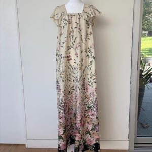 May include: A cream-colored, floor-length dress with a square neckline and short, ruffled sleeves. The dress is adorned with a floral pattern in shades of pink, purple, and green, with a black border at the hem. The dress is displayed on a mannequin.