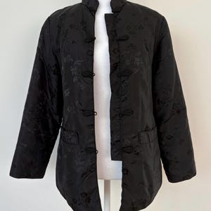May include: Black quilted jacket with a mandarin collar and frog closures. The jacket features a floral pattern and two front pockets. The sleeves are long and the jacket is open in the front.
