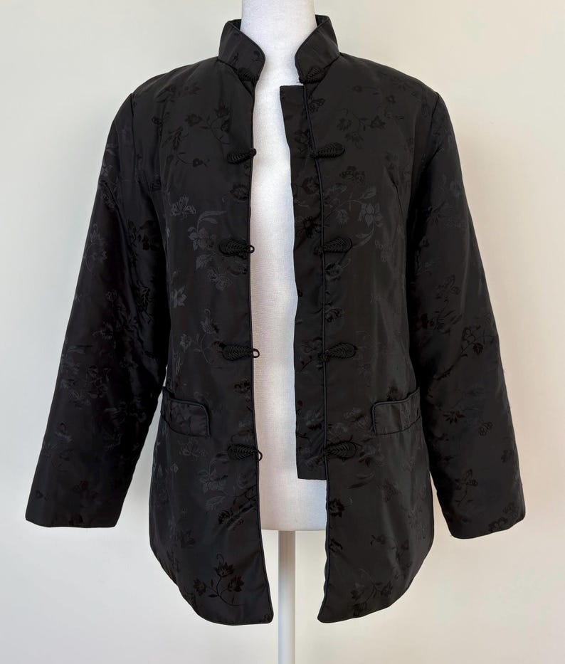 May include: Black quilted jacket with a mandarin collar and frog closures. The jacket features a floral pattern and two front pockets. The sleeves are long and the jacket is open in the front.