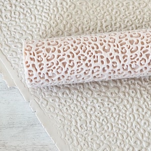 May include: A close-up of a pink and white rolling pin with a leopard print design. The rolling pin is lying on a sheet of white fondant with a similar leopard print design.