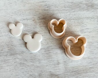 May include: Two white fondant Mickey Mouse head shapes and two plastic Mickey Mouse head cookie cutters on a white wooden surface.