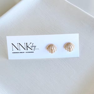 May include: A pair of gold-colored seashell stud earrings on a white card. The card has the text "NNK clay co." and "HANDMADE JEWELRY + ACCESSORIES" printed in black. The earrings are displayed on a white surface.