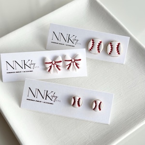 May include: Three pairs of handmade earrings on white cards. One pair features white bows with red accents, and two pairs are designed as baseballs with red stitching. The cards display the text "NNK clay co. HANDMADE JEWELRY + ACCESSORIES."