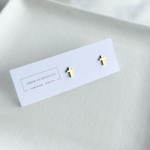 May include: Gold-colored cross stud earrings on a white card with the text "URBAN VA DESIGN CO HANDMADE JEWELRY". The earrings are small and minimalist, perfect for everyday wear.