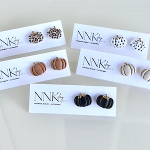 May include: A collection of pumpkin-shaped stud earrings in various colors and patterns, including leopard print, polka dots, brown, white, and black. Each pair is displayed on a white card with the text "NNK clay" and "HANDMADE JEWELRY + ACCESSORIES".
