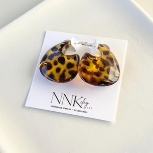 May include: A pair of teardrop-shaped earrings with a tortoiseshell pattern. The earrings are brown and yellow with dark brown spots. The earrings are displayed on a white card with the text "NNKclay co. HANDMADE JEWELRY + ACCESSORIES."