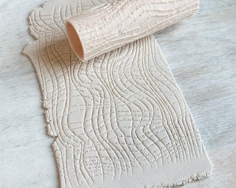 May include: A light beige clay roller and two clay sheets with a wood grain texture. The cylindrical roller is positioned on top of one of the sheets. The clay sheets have a rectangular shape with irregular edges.