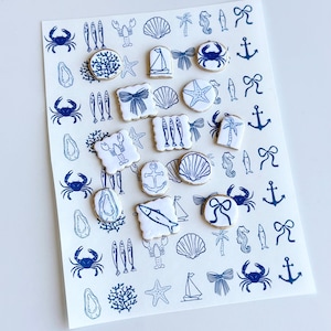 May include: Sheet of nautical-themed stickers with blue illustrations on a white background. Designs include crabs, fish, seashells, starfish, sailboats, bows, anchors, seahorses, and palm trees. The stickers are arranged in a grid pattern.