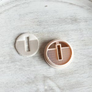May include: Two round, beige plastic cookie cutters. The larger cutter has a design of vertical lines with a vertical rectangle in the center. The smaller cutter has a design of vertical lines.