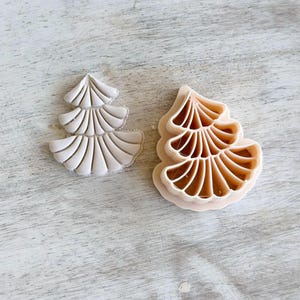May include: Two plastic Christmas tree-shaped cookie cutters. One is a solid off-white color, and the other is a light tan color. The cutters have a layered design with fan-like details, perfect for holiday baking.