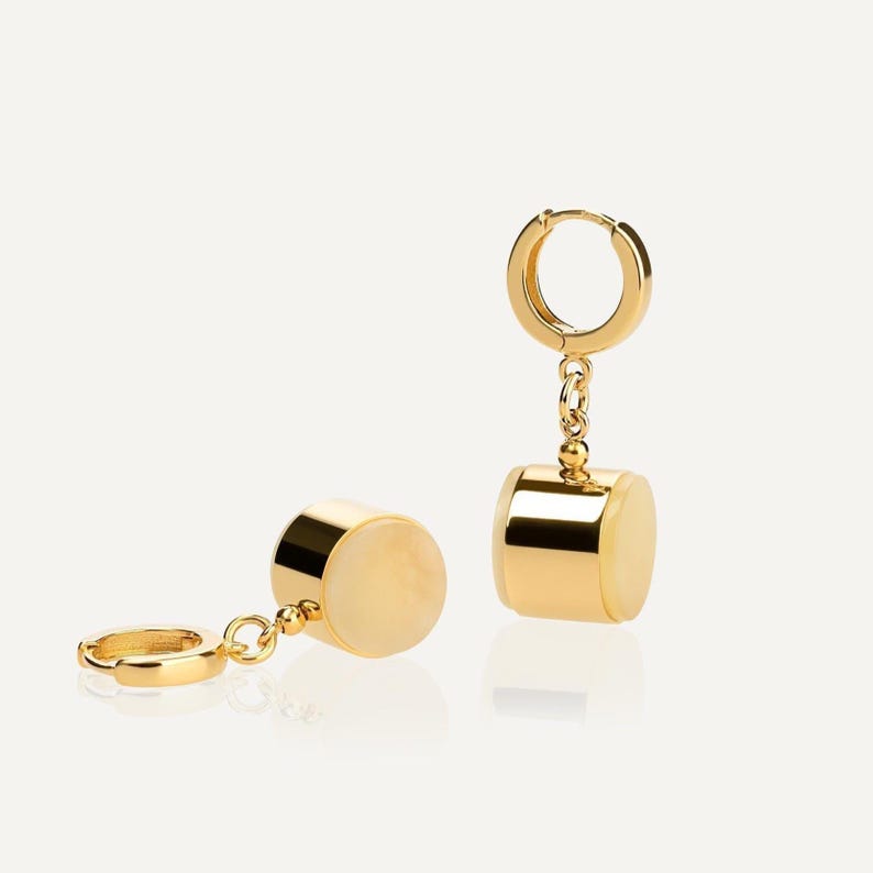 May include: Two gold-colored hoop earrings with a cylindrical, off-white stone pendant. The earrings are displayed against a white background, showcasing their polished, reflective surface and elegant design.