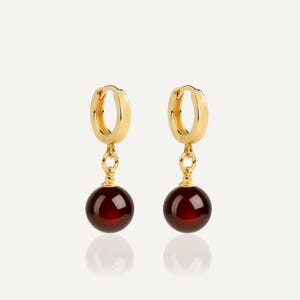 May include: A pair of gold hoop earrings featuring a polished finish, each suspending a single, smooth, spherical dark red gemstone bead. The earrings are designed with a hinged closure mechanism, showcasing a classic drop style with vibrant red accents.