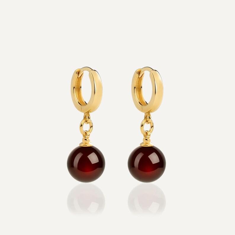 May include: A pair of gold hoop earrings featuring a polished finish, each suspending a single, smooth, spherical dark red gemstone bead. The earrings are designed with a hinged closure mechanism, showcasing a classic drop style with vibrant red accents.