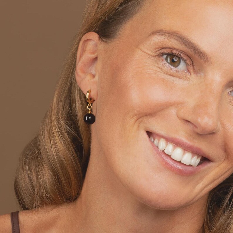 May include: A gold hoop earring featuring a small, smooth, spherical black bead charm suspended from the bottom. The earring is crafted from a polished gold-tone metal, creating a simple and classic design that hangs vertically from the earlobe.