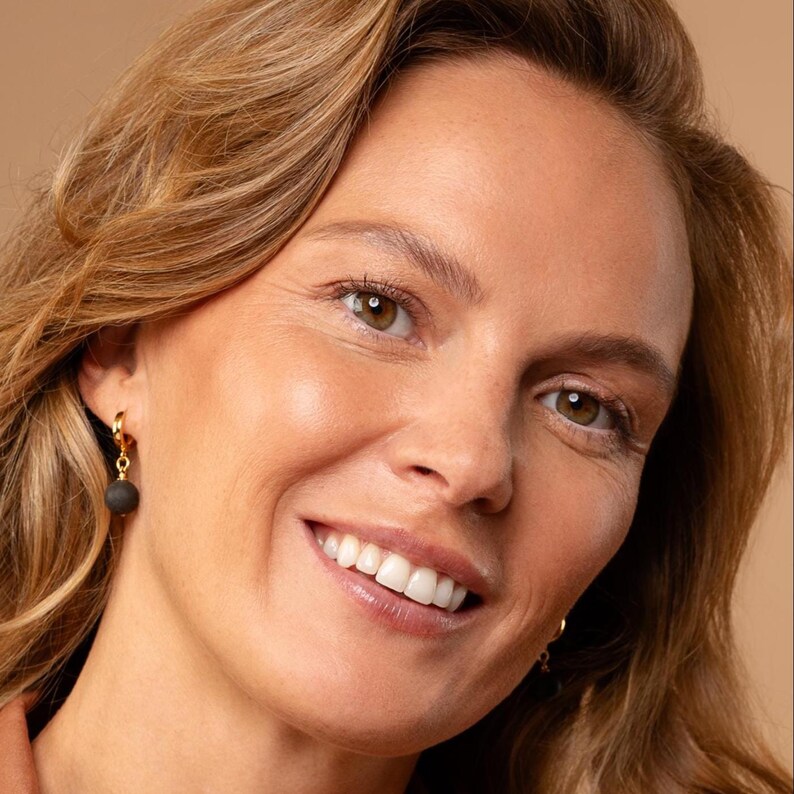 May include: A gold hoop earring featuring a small, matte black spherical bead charm. The earring is constructed with a polished gold-tone metal hoop that connects to a dark, textured round bead, creating a simple and contrasting jewelry piece.