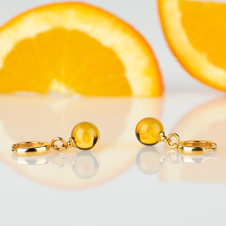 May include: A pair of gold hoop earrings with amber-colored round beads hanging from them. The earrings are on a white surface.