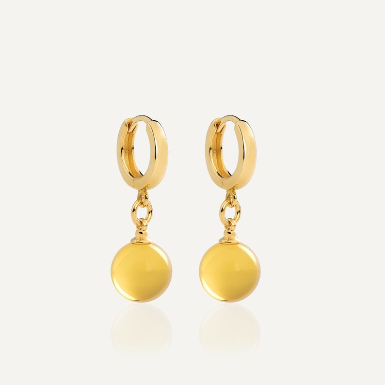 May include: A pair of gold hoop earrings with a dangling yellow orb charm. The earrings are simple and elegant.