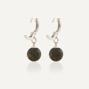 May include: A pair of silver hoop earrings featuring a hinged closure and a dangling spherical charm. Each charm is a dark, mottled brown stone bead with a smooth, polished finish. The silver hoops are stamped with 925, indicating sterling silver.