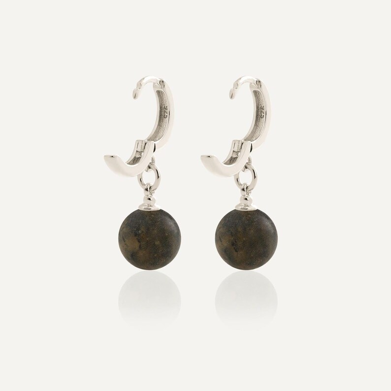 May include: A pair of silver hoop earrings featuring a hinged closure and a dangling spherical charm. Each charm is a dark, mottled brown stone bead with a smooth, polished finish. The silver hoops are stamped with 925, indicating sterling silver.