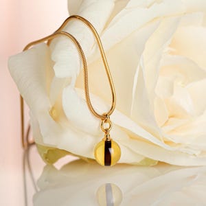 May include: A gold necklace with a yellow pendant and a dark brown stripe. The necklace is set against a white rose. The pendant is reflected on a shiny surface below.